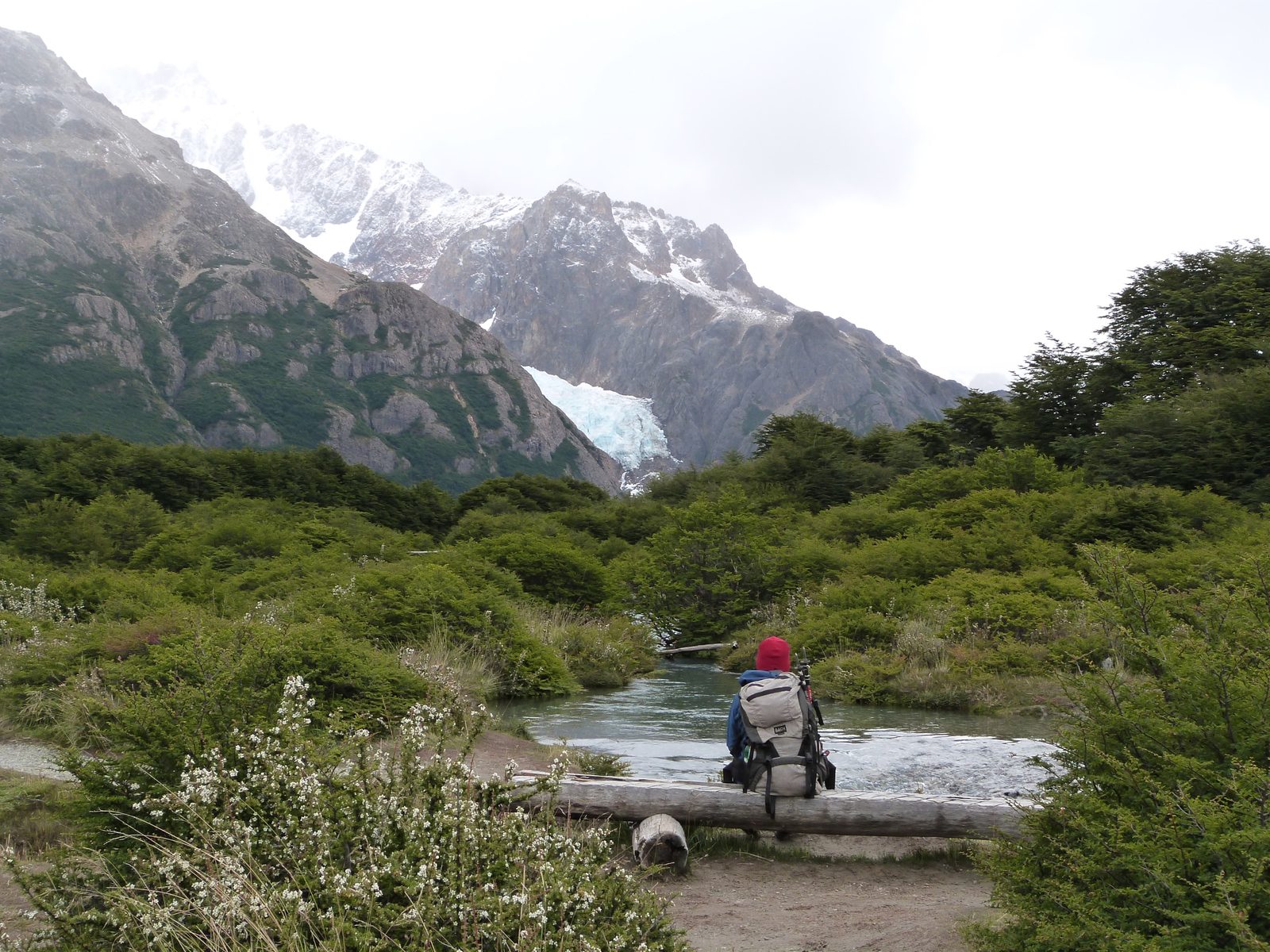 Caminante de gorro rojo sentado sobre un tronco, de espaldas, mirando el glaciar